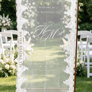K&ouml;nnte beinhalten: Ein hohes, rechteckiges, durchscheinendes Acryl-Hochzeitsschild mit goldfarbenem Metallrahmen und wei&szlig;er Spitzenborte an den vertikalen Kanten. Der Text lautet: Welcome to THE WEDDING OF, das Monogramm HM, ALL THINGS SALAM, HEIDI & MAXWELL, 06.02.2028.
