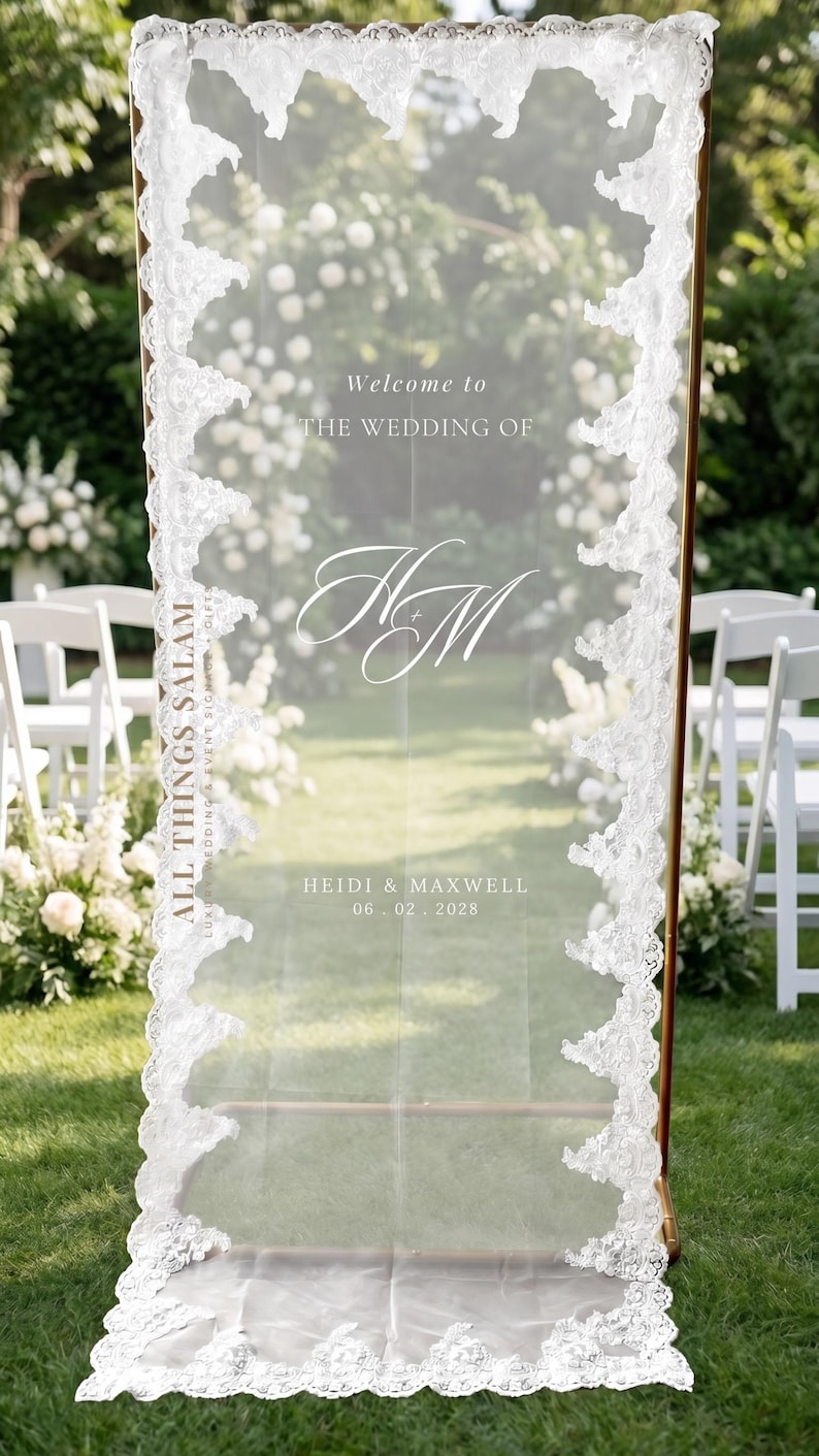 K&ouml;nnte beinhalten: Ein hohes, rechteckiges, durchscheinendes Acryl-Hochzeitsschild mit goldfarbenem Metallrahmen und wei&szlig;er Spitzenborte an den vertikalen Kanten. Der Text lautet: Welcome to THE WEDDING OF, das Monogramm HM, ALL THINGS SALAM, HEIDI & MAXWELL, 06.02.2028.