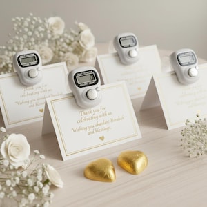 May include: White tally counters with digital displays are clipped to folded cards with gold trim and text. The cards read "Thank you for celebrating with us. Wishing you abundant Barakah and blessings." Gold foil heart-shaped candies are at the bottom.