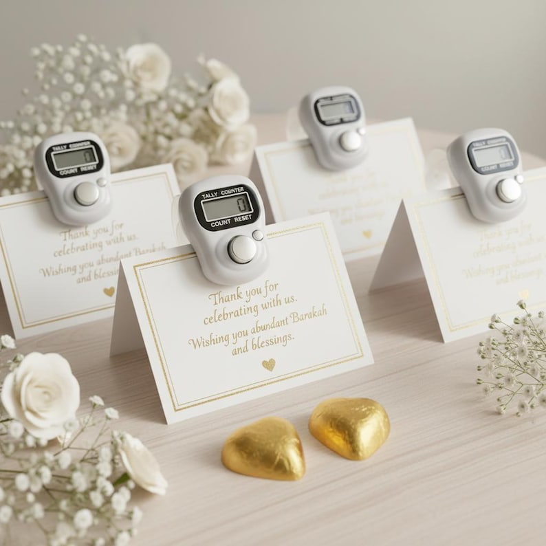 May include: White tally counters with digital displays are clipped to folded cards with gold trim and text. The cards read "Thank you for celebrating with us. Wishing you abundant Barakah and blessings." Gold foil heart-shaped candies are at the bottom.