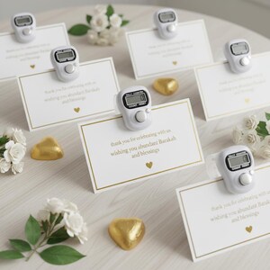 May include: White cards with gold borders and text, topped with white tally counters. The cards read "thank you for celebrating with us wishing you abundant Barakah and blessings." Gold heart-shaped candies and white flowers are scattered around.