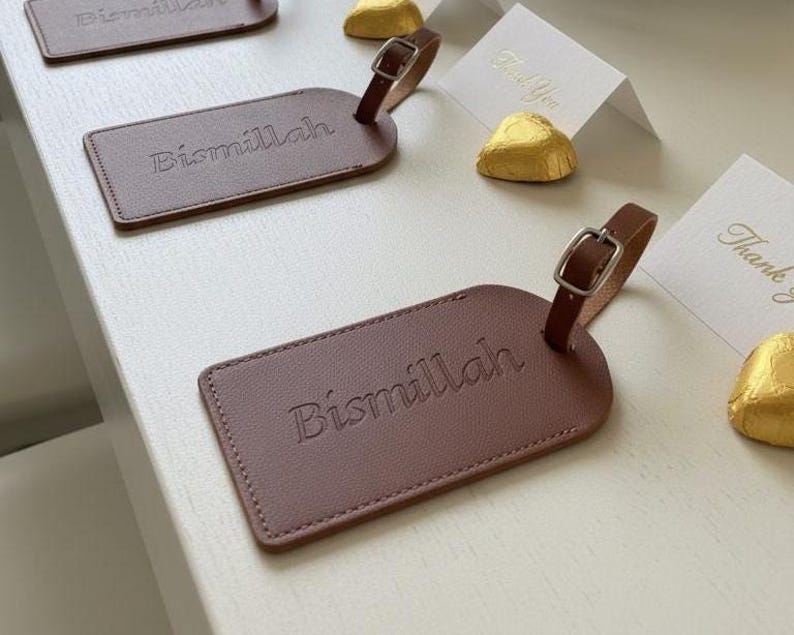 Pu&ograve; includere: Etichette per bagagli in pelle marrone con la scritta "Bismillah" in rilievo sul davanti. Ogni etichetta ha un cinturino in pelle marrone con fibbia argentata. Sono visibili anche biglietti bianchi con testo dorato "Thank You".