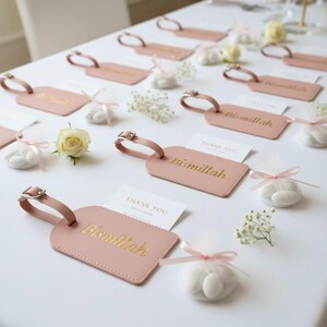 May include: Pink luggage tags with gold lettering that reads "Bismillah" are arranged on a white surface. Small white candies are tied with pink ribbons. White flowers and "Thank You" cards are also present.