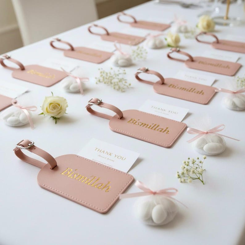 May include: Pink luggage tags with gold lettering that reads "Bismillah" are arranged on a white surface. Small white candies are tied with pink ribbons. White flowers and "Thank You" cards are also present.