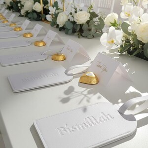 Pu&ograve; includere: Etichette per bagagli in pelle bianca con la scritta in rilievo "Bismillah" sono esposte su una superficie bianca. Sono presenti anche piccoli biglietti bianchi con "Thank You" in lettere dorate e cioccolatini dorati. Composizioni floreali con rose e orchidee bianche.
