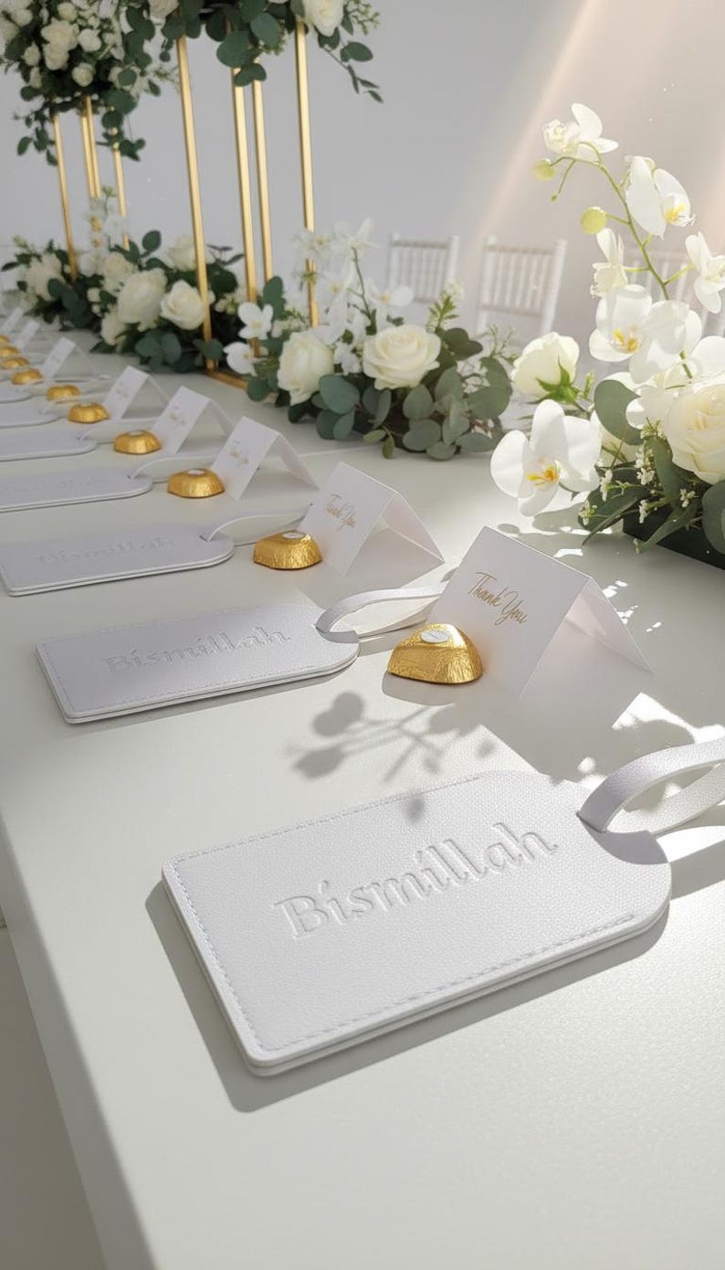 Pu&ograve; includere: Etichette per bagagli in pelle bianca con la scritta in rilievo "Bismillah" sono esposte su una superficie bianca. Sono presenti anche piccoli biglietti bianchi con "Thank You" in lettere dorate e cioccolatini dorati. Composizioni floreali con rose e orchidee bianche.