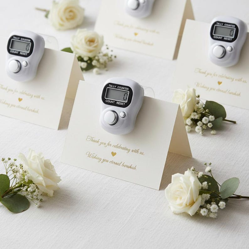 May include: White tally counters with digital displays are placed on folded cards with gold text and a small heart. The cards are surrounded by white roses and greenery. The text on the cards reads, "Thank you for celebrating with us. Wishing you eternal barakah."