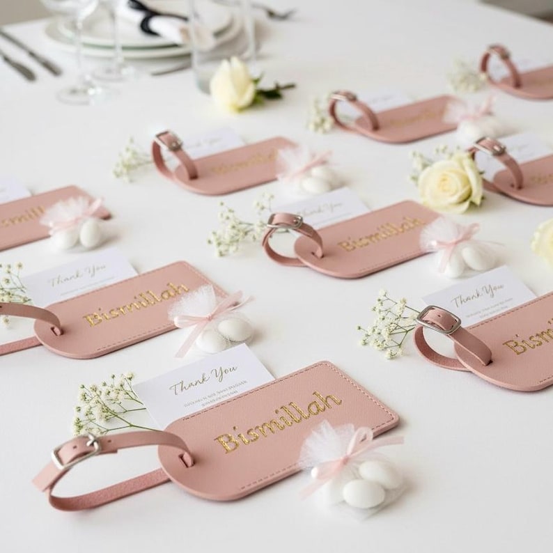 May include: Pink luggage tags with gold lettering that reads "Bismillah" are arranged on a white surface. Each tag has a buckle and is paired with a small card that says "Thank You". Small white candies are also present.