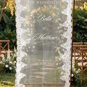 K&ouml;nnte beinhalten: Ein hohes, rechteckiges, transparentes Acryl-Willkommensschild f&uuml;r Hochzeiten mit einem goldfarbenen Metallrahmen. Es tr&auml;gt den wei&szlig;en Schriftzug "welcome to THE WEDDING OF Bella & Matthew 06.08.2030 THE LUSH OASIS" und ist mit wei&szlig;er Spitze verziert.