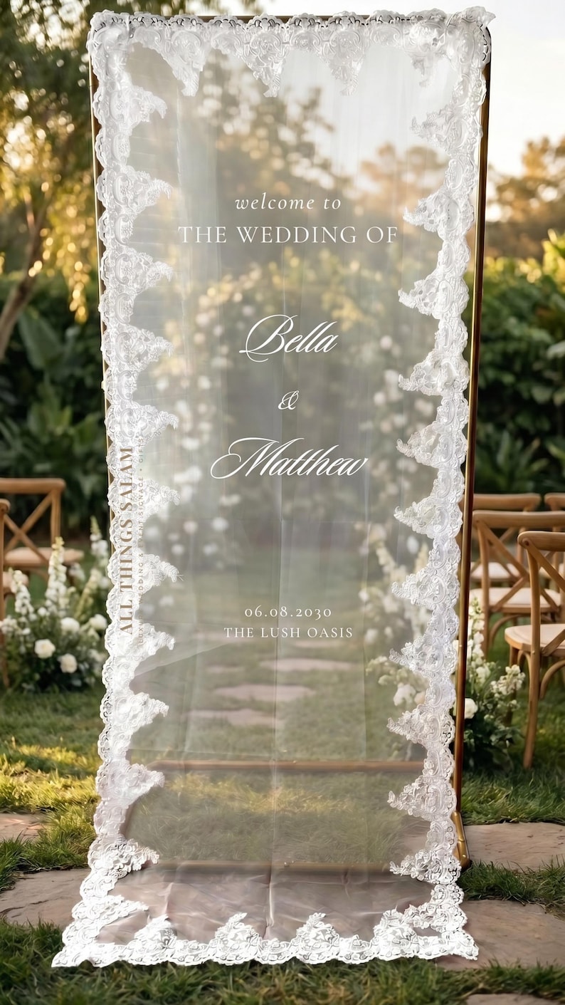 K&ouml;nnte beinhalten: Ein hohes, rechteckiges, transparentes Acryl-Willkommensschild f&uuml;r Hochzeiten mit einem goldfarbenen Metallrahmen. Es tr&auml;gt den wei&szlig;en Schriftzug "welcome to THE WEDDING OF Bella & Matthew 06.08.2030 THE LUSH OASIS" und ist mit wei&szlig;er Spitze verziert.