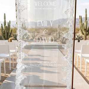 K&ouml;nnte beinhalten: Ein rechteckiges Willkommensschild f&uuml;r Hochzeiten aus klarem Acryl mit goldfarbenem Metallrahmen, verziert mit wei&szlig;er Spitzenborte an den Seiten. Der Text lautet: WELCOME TO THE WEDDING OF EMILY & DAVID, MAY 28TH, 2027, PALM SPRINGS, CALIFORNIA, und ALL THINGS SALAM.