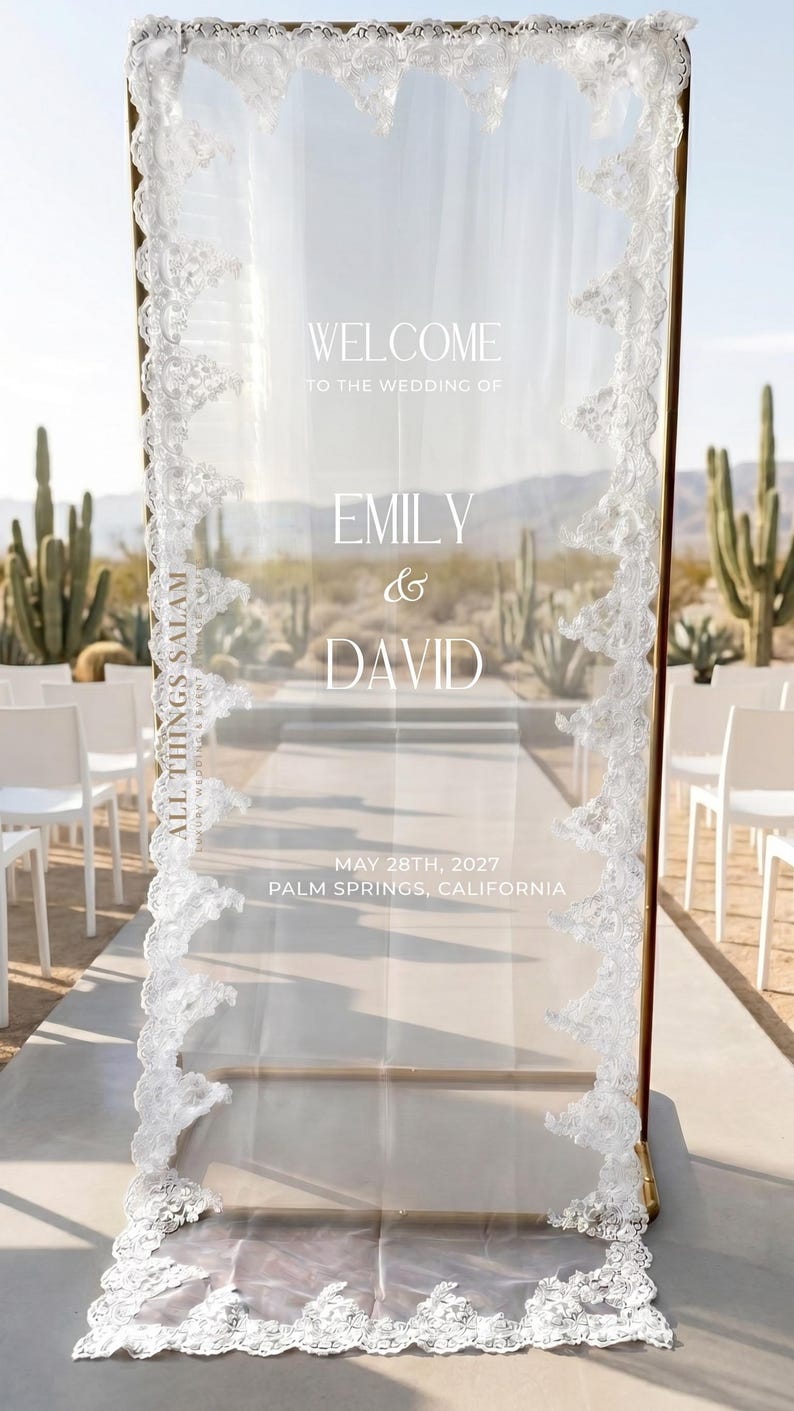 K&ouml;nnte beinhalten: Ein rechteckiges Willkommensschild f&uuml;r Hochzeiten aus klarem Acryl mit goldfarbenem Metallrahmen, verziert mit wei&szlig;er Spitzenborte an den Seiten. Der Text lautet: WELCOME TO THE WEDDING OF EMILY & DAVID, MAY 28TH, 2027, PALM SPRINGS, CALIFORNIA, und ALL THINGS SALAM.