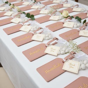 May include: Arrangement of pink luggage tags with gold lettering that reads "Bismillah" and small white cards that say "Thank You". Each tag has a matching strap and is paired with a small bag of white candies and floral accents.
