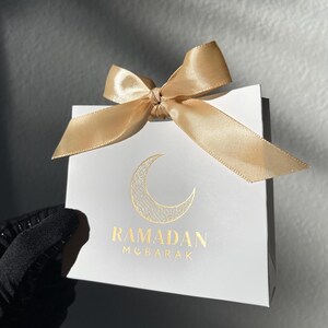 May include: White gift bag with a gold satin bow. The bag features a gold crescent moon design and the words "RAMADAN MUBARAK" in gold lettering. The bag is held by a gloved hand.