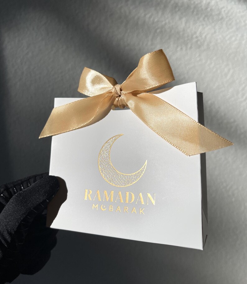 May include: White gift bag with a gold satin bow. The bag features a gold crescent moon design and the words "RAMADAN MUBARAK" in gold lettering. The bag is held by a gloved hand.