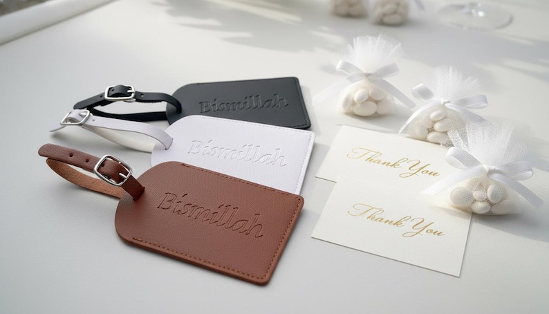 Pu&ograve; includere: Etichette per bagagli in pelle nera, bianca e marrone, ciascuna con fibbia e la parola "Bismillah" in rilievo. Accanto alle etichette, piccoli biglietti bianchi con "Thank You" in corsivo dorato e piccoli sacchetti di caramelle bianche.