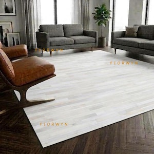 May include: A large rectangular area rug featuring a patchwork design of light gray and white rectangular strips. The rug is positioned on a dark brown herringbone wood floor in a room with gray sofas and a brown leather chair. Text reads FIORWYN.