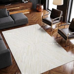 Op de afbeelding: Een rechthoekig, gebroken wit tapijt met een geometrisch patroon van diagonale lijnen. Het tapijt ligt op een donkere houten vloer en de kamer heeft een donkergrijze bank, twee leren stoelen en een vloerlamp. De tekst "FIORWYN" is zichtbaar op het tapijt.