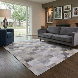 May include: A rectangular area rug featuring a patchwork design of square tiles in varying shades of gray, silver, and white. The rug is positioned on a dark wood floor in a living room with a gray sofa, a blue armchair, and a floor lamp.