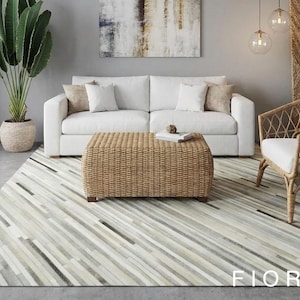 Può includere: Un soggiorno moderno con un divano bianco, un pouf intrecciato e una sedia in rattan. Un tappeto a righe in tonalità neutre copre il pavimento. Una grande pianta e opere d'arte completano l'arredamento. La parola "FIORWYN" è visibile sul tappeto.
