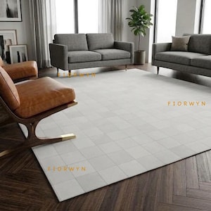 May include: A large rectangular area rug featuring a light grey grid pattern of square tiles. The rug is positioned on a dark brown herringbone wood floor in a room with grey sofas, a brown leather chair, and the text FIORWYN visible on it.