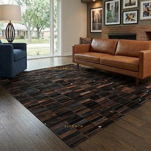 May include: A rectangular area rug featuring a patchwork design of dark brown, black, and tan leather tiles. The rug is positioned on a dark wood floor in a living room, extending from beneath a tan leather sofa toward a blue armchair.
