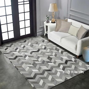 Puede incluir: Una alfombra rectangular con un patrón de chevrón en tonos grises, blancos y negros. La alfombra está colocada sobre un suelo de hormigón frente a un sofá blanco. El texto "FIORWYN" es visible en la alfombra.