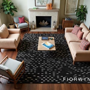 Op de afbeelding: Een woonkamer met een zwart-wit patroon tapijt, twee beige banken en twee fauteuils. Een houten salontafel staat in het midden en een open haard bevindt zich op de achtergrond. Het woord "FIORWYN" is zichtbaar op het tapijt.