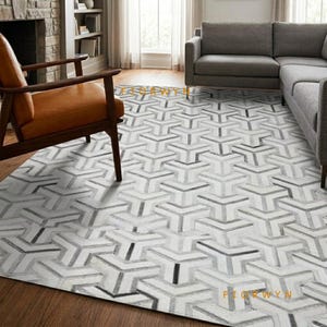Könnte beinhalten: Ein rechteckiger Teppich mit einem sich wiederholenden geometrischen Muster aus ineinandergreifenden Y-förmigen Segmenten in Weiß, Grau und Schwarz. Der Teppich liegt auf einem Holzboden unter einem braunen Ledersessel und einem grauen Stoffsofa.