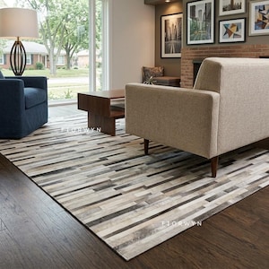 Peut inclure: Un tapis rectangulaire présentant un motif en patchwork composé de bandes horizontales dans des tons variés de gris, blanc et marron. Le tapis est placé sur un parquet sombre sous un canapé beige, une table basse en bois et un fauteuil bleu.