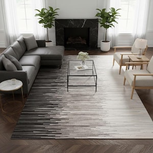 May include: A contemporary living room with a grey corner sofa, two armchairs, and a glass coffee table. A large rug with a black and white linear pattern is on the floor. Two potted plants are either side of a black marble fireplace.