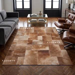 Op de afbeelding: Een rechthoekig patchwork tapijt in bruin en beige met het woord FIORWYN erop gedrukt. Het tapijt is gemaakt van rechthoekige en vierkante stukken huid, aan elkaar gestikt met zichtbare klinknagels. Het tapijt ligt op een donkere houten vloer.