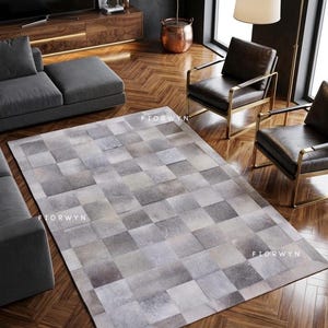 May include: A rectangular area rug with a patchwork design in shades of gray and white. The rug is made of squares of varying tones, creating a textured appearance. The rug is placed on a hardwood floor in a living room setting.