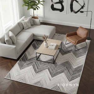 Peut inclure: Un salon moderne avec un canapé d'angle gris clair, un fauteuil en cuir marron et une table basse en bois. Un grand tapis à motifs chevrons noirs et blancs recouvre le sol. Deux peintures abstraites en noir et blanc ornent le mur.