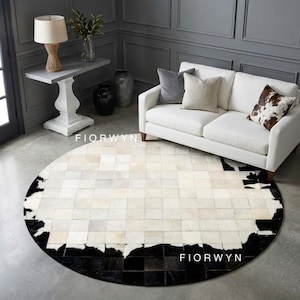 May include: A round cowhide rug with a patchwork design. The rug features a black border and a central pattern of white and cream-colored squares. The rug is placed in a living room setting with a white sofa and gray walls.
