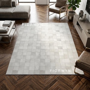Puede incluir: Una alfombra grande, rectangular, de color gris claro con un patrón de cuadrícula. La alfombra está hecha de cuadrados de diferentes tonos de gris. El nombre de la marca "FIORWYN" está impreso en la alfombra. La alfombra está colocada sobre un suelo de madera oscura.