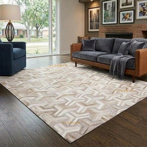 Peut inclure: Un grand tapis rectangulaire présentant un motif géométrique répétitif dans des tons beige, crème et gris clair. Le tapis est placé sur un parquet en bois foncé dans un salon, s'étendant du premier plan vers un canapé marron.