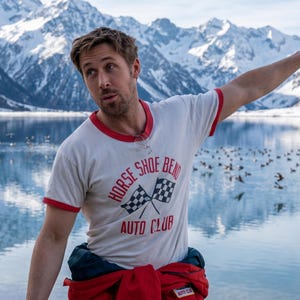 May include: A white t-shirt with red trim featuring the text "HORSE SHOE BEND AUTO CLUB" and two checkered flags. The person is standing near a lake with mountains in the background.