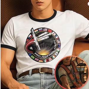 May include: White t-shirt with black trim displaying a circular graphic with a spaceship design, the text "PROJECT HAIL MARY," and flags of various nations. The shirt is worn with blue jeans and a brown belt.
