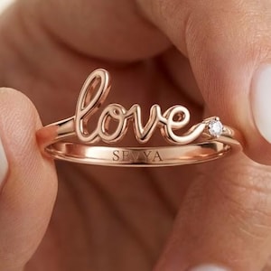 Könnte beinhalten: Ein Ring aus Roségold mit dem Wort love in Schreibschrift als Herzstück. Das Band enthält einen kleinen, klaren, rund geschliffenen Edelstein rechts neben dem Schriftzug und das Wort SEVYA ist auf der Innenseite des Rings eingraviert.