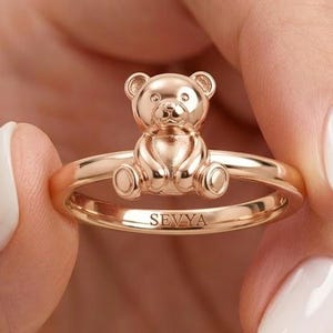 Puede incluir: Un anillo de oro rosa que presenta una pequeña figura de oso de peluche tridimensional centrada en la banda. El interior de la banda del anillo está grabado con el texto SEVYA en letras mayúsculas. El metal tiene un acabado pulido.