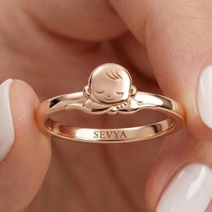 Könnte beinhalten: Ein Ring aus Roségold mit einem skulpturalen Babygesicht-Design auf der Ringschiene. Die Innenseite der Schiene ist mit dem Namen SEVYA in Großbuchstaben graviert. Der Ring hat eine polierte metallische Oberfläche und eine glatte Form.