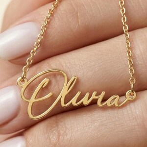 Könnte beinhalten: Eine goldfarbene Halskette mit einem Namensanhänger in Schreibschrift, der den Namen Olivia zeigt. Der Anhänger ist an einer zarten, dünnen Gliederkette befestigt. Das Metall hat eine polierte Oberfläche und die Buchstaben sind verbunden.