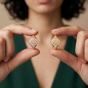 Könnte beinhalten: Zwei ovale Siegelringe aus poliertem Gold, jeweils mit einer zentralen ovalen Platte, die von einem Rand aus kleinen, klaren, rund geschliffenen Steinen umgeben ist. Der linke Ring hat eine schlichte Mitte, der rechte eine massive Goldmitte.
