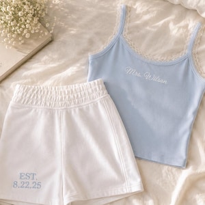May include: A light blue camisole with white lace trim and the embroidered text Mrs. Wilson, paired with white shorts featuring an elastic waistband and the embroidered blue text EST. 8.22.25. The set is displayed on a soft, textured fabric surface.