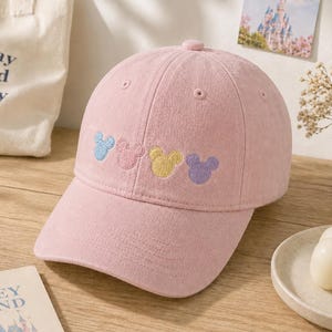 Op de afbeelding: Lichtroze baseballpet met een gebogen klep. De pet heeft geborduurde Mickey Mouse-hoofdontwerpen in blauw, roze, geel en paars. Bovenop zit een kleine, bijpassende roze knoop. Een canvas draagtas en een ansichtkaart staan op de achtergrond.