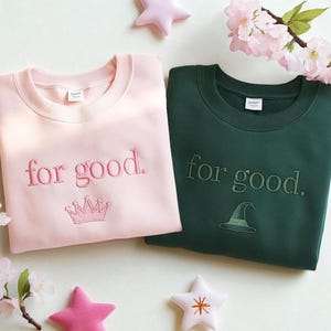 May include: Two crewneck sweatshirts, one light pink and one dark green, embroidered with "for good." The pink sweatshirt features a crown, and the green one has a witch hat. Decorative stars and flowers surround the sweatshirts.