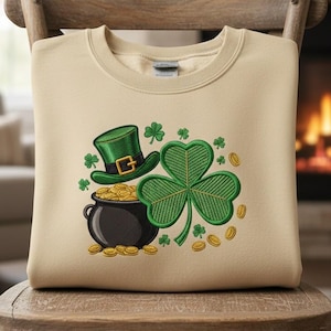 May include: Beige sweatshirt featuring a St. Patrick's Day design. The design includes a green leprechaun hat, a pot of gold, a large green shamrock, and gold coins. The sweatshirt is displayed on a wooden chair.