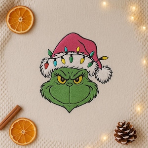 Könnte beinhalten: Gesticktes Bild einer grünen Figur mit einer rosa Weihnachtsmütze, die mit bunten Lichterketten geschmückt ist. Die Figur hat einen grimmigen Gesichtsausdruck mit gelben Augen. Das Bild befindet sich auf einem cremefarbenen Hintergrund mit Orangenscheiben, Zimtstange und Tannenzapfen.