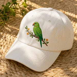 Puede incluir: Una gorra de béisbol blanca con un diseño bordado de un loro verde brillante posado sobre una rama marrón con pequeñas flores blancas y amarillas. La gorra tiene una corona estructurada, una visera curva y ojales cosidos en la parte superior.